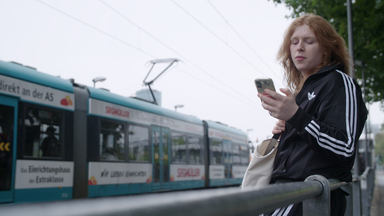 Yuna steht an einer Haltestelle. Im Hintergrund fährt eine Straßenbahn.