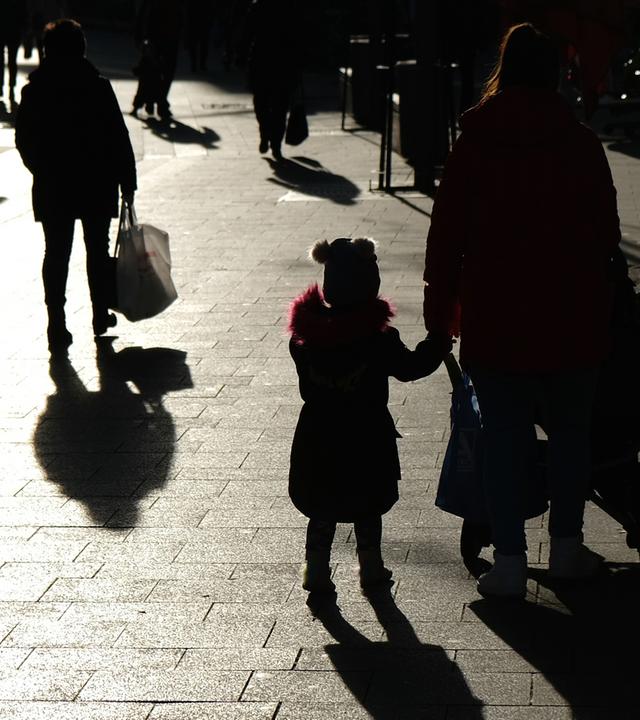Passanten mit Tüten gehen während des Lockdowns durch eine Fußgängerzone und sind dabei als Silhouette und Schatten zu sehen.