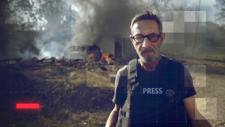 frontal-Reporter Arndt Ginzel mit schusssicherer Weste vor brennendem Autofrack, das durch einen Drohnenangriff in der Ukraine zerstört wurde 