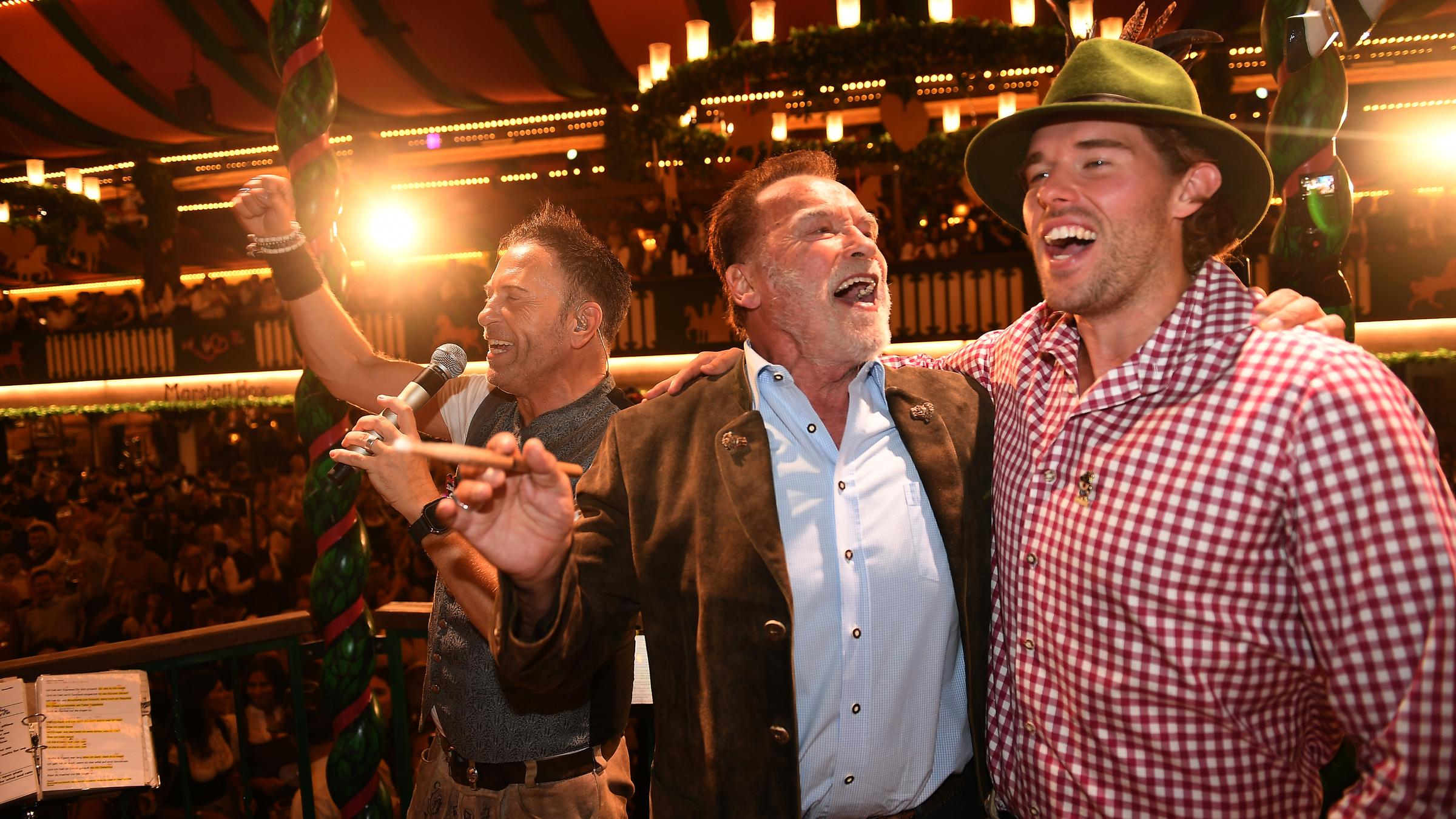 26.09.2025, Bayern, München: Arnold Schwarzenegger und sein Sohn Christopher Schwarzenegger stehen im Marstall-Festzelt auf der Bühne und dirigieren.
