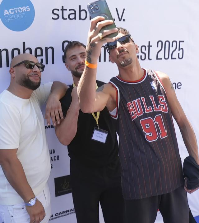 Drei junge Männer mit Kinnbärten stehen vor einer Logowand vom Münchner Filmfest. Einer hält sein Handy für ein Selfie hoch.