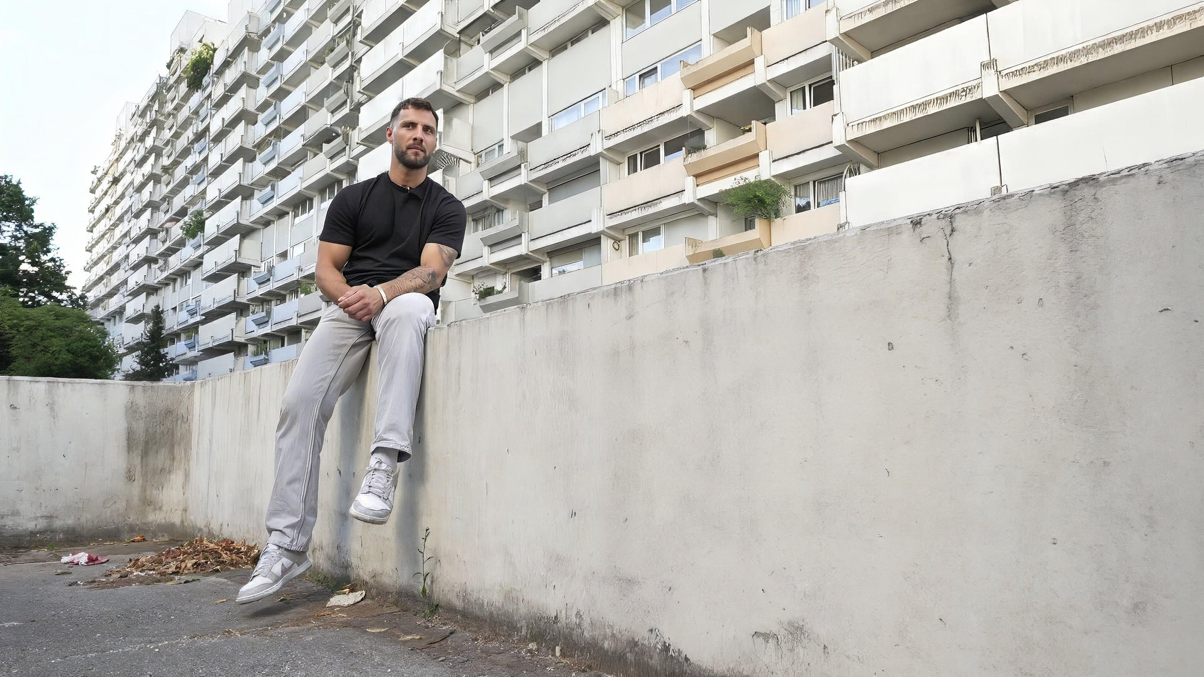 Ein junger Mann sitzt auf einer Betonwand vor einem Hochhaus