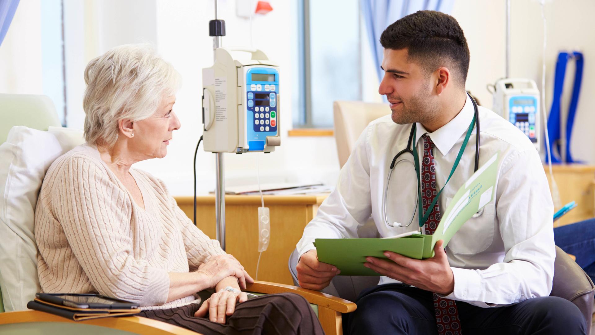 Eine Patientin erhält eine Chemotherapie per Infusion. Ein Arzt sitzt ihr im Gespräch gegenüber.