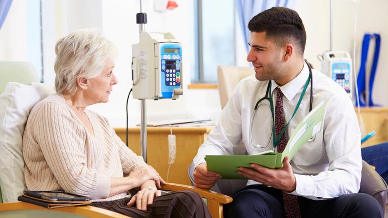 Eine Patientin erhält eine Chemotherapie per Infusion. Ein Arzt sitzt ihr im Gespräch gegenüber.