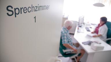 Ein älterer Mann mit weißen Haaren sitzt bei einer Ärztin im Sprechzimmer am Schreibtisch.