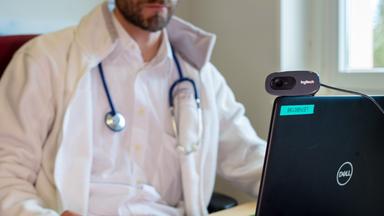 symbolbild:  ein hausarzt spricht waehrend einer videosprechstunde in seiner praxis mit einer patientin. 