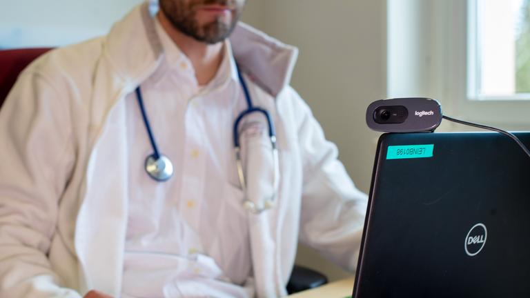 symbolbild:  ein hausarzt spricht waehrend einer videosprechstunde in seiner praxis mit einer patientin. 