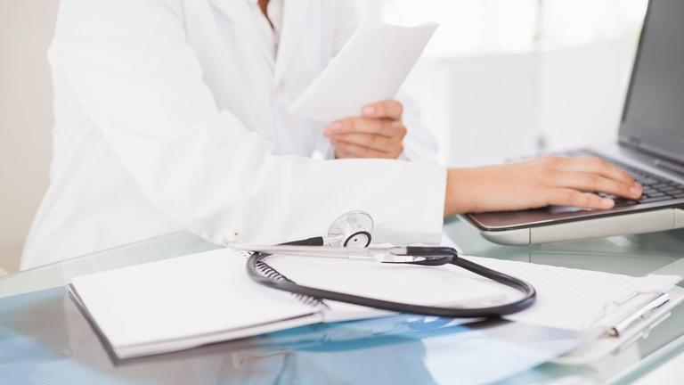 Ein Arzt sitzt in einer Arztpraxis hinter einem Glastisch. Auf diesem liegen Papierunterlagen und darauf ein Stethoskop. Der Mediziner tippt auf einem Laptop