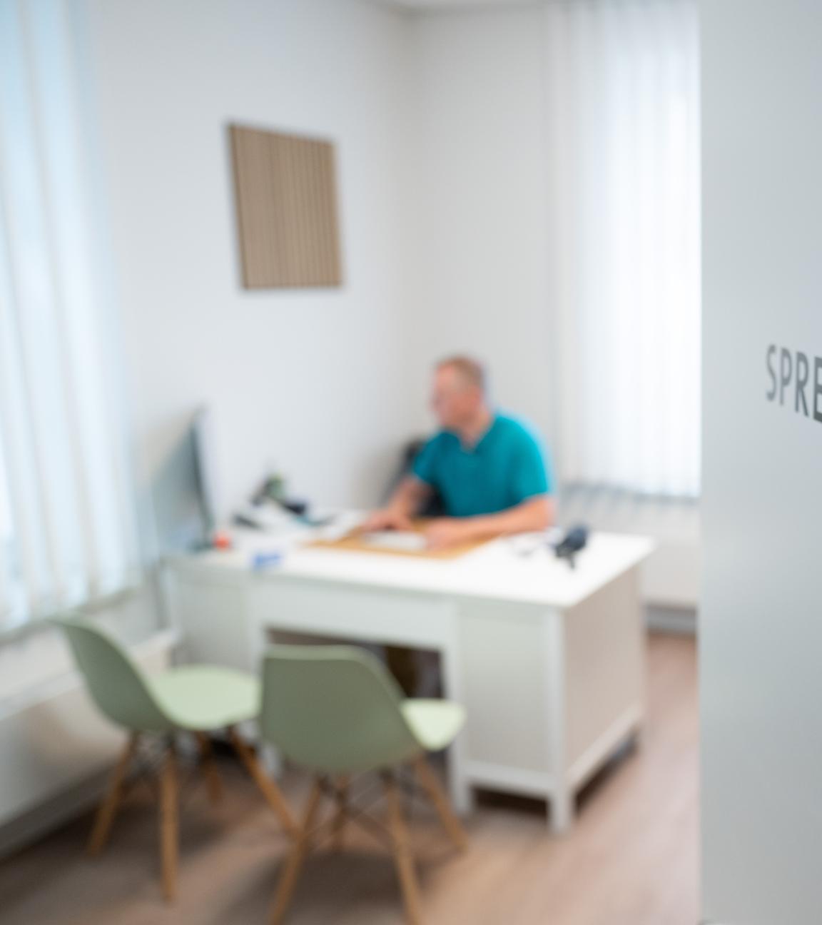 Ein Arzt sitzt in einem Sprechzimmer seiner Hausarztpraxis an einem Schreibtisch und arbeitet am Computer. 