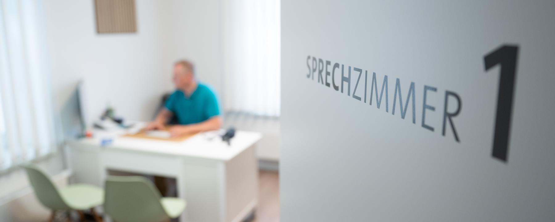 Ein Arzt sitzt in einem Sprechzimmer seiner Hausarztpraxis an einem Schreibtisch und arbeitet am Computer. 