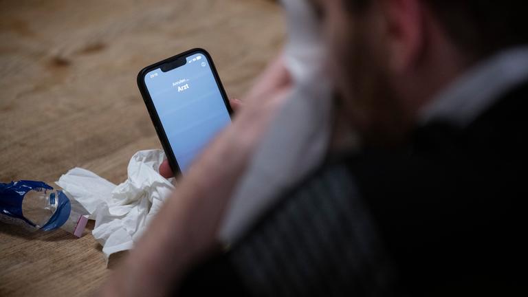 Ein Mann hält ein Taschentuch in einer Hand und ein Telefon mit dem Schriftzug «Arzt» in der Anderen.
