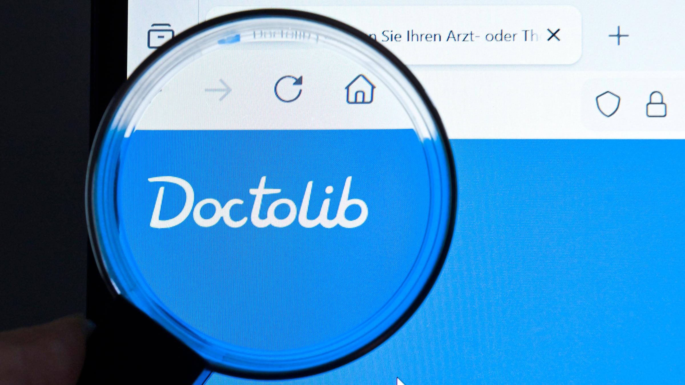 Website des Patientenportals Doctolib durch eine Lupe gesehen, die vor einen Monitor gehalten wird.