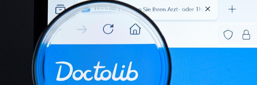 Website des Patientenportals Doctolib durch eine Lupe gesehen, die vor einen Monitor gehalten wird.