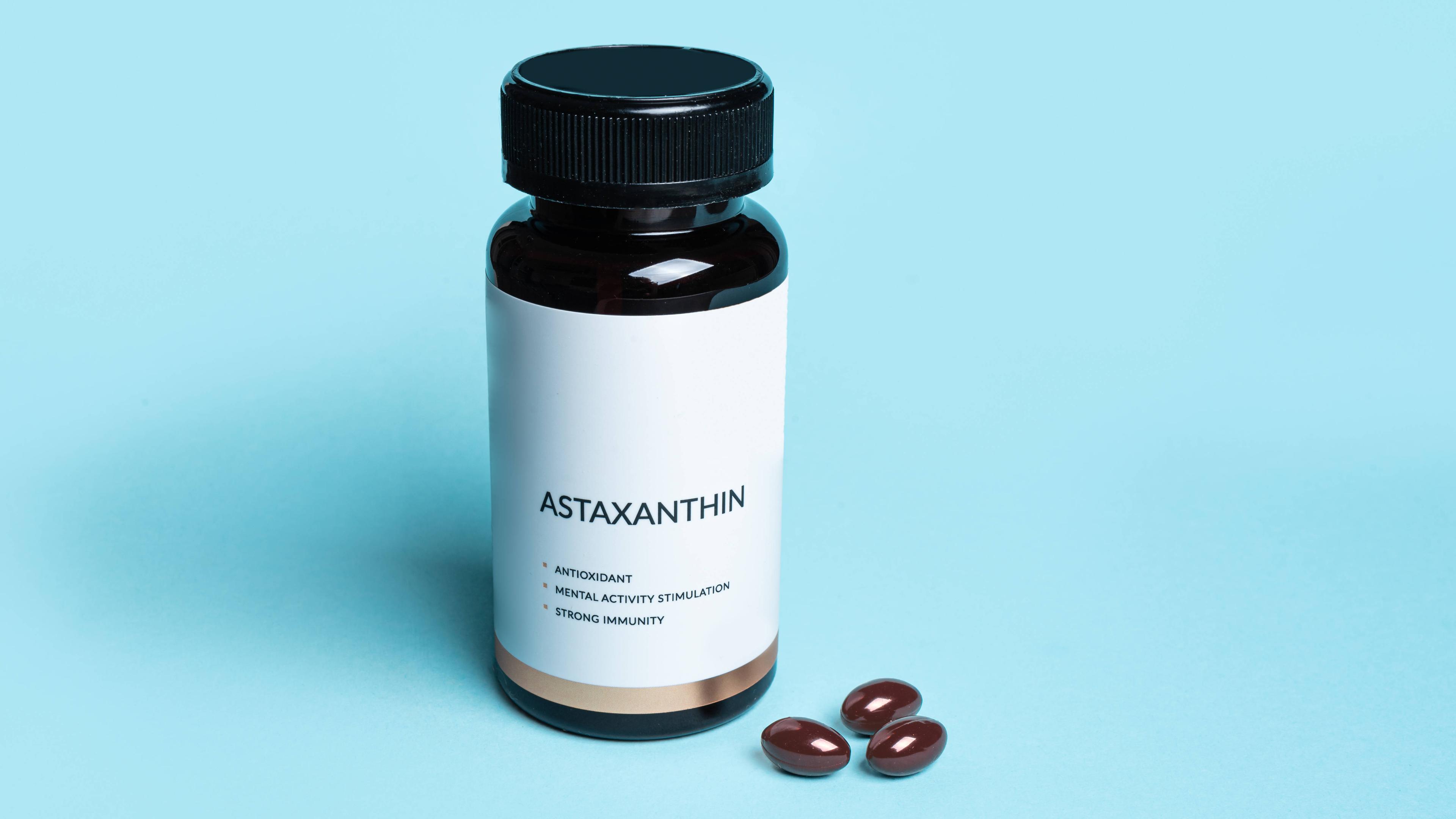 Eine braune Flasche Astaxanthin mit der Aufschrift "ASTAXANTHIN / ANTIOXIDANT / MENTAL ACITVITY STIMULATION / STRONG IMMUNITY" vor einem blauen Hintergrund, daneben drei Kapseln des Nahrungsergänzungsmittels.