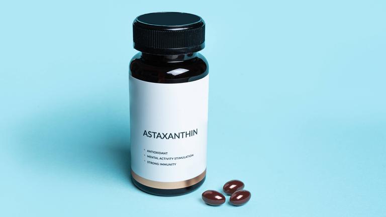 Eine braune Flasche Astaxanthin mit der Aufschrift "ASTAXANTHIN / ANTIOXIDANT / MENTAL ACITVITY STIMULATION / STRONG IMMUNITY" vor einem blauen Hintergrund, daneben drei Kapseln des Nahrungsergänzungsmittels.