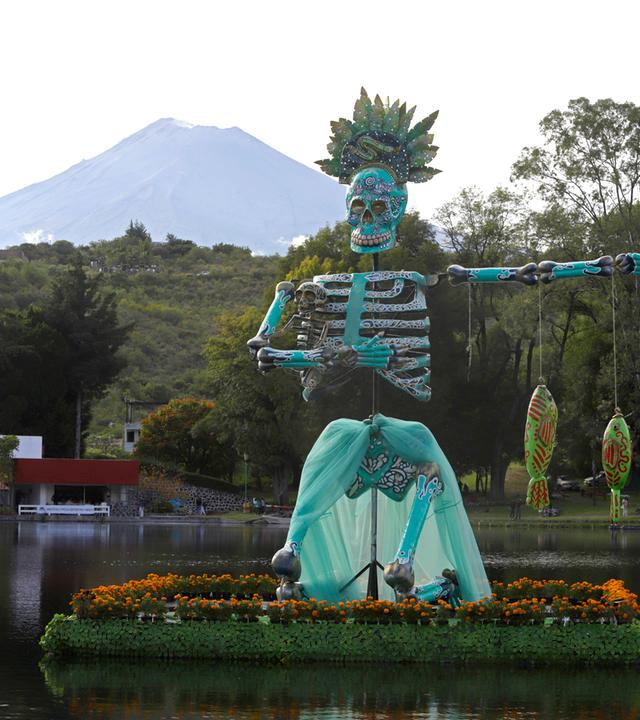 Ein bunt bemaltes Skelett steht auf einem See. Im Hintergrund der Popocatépetl.