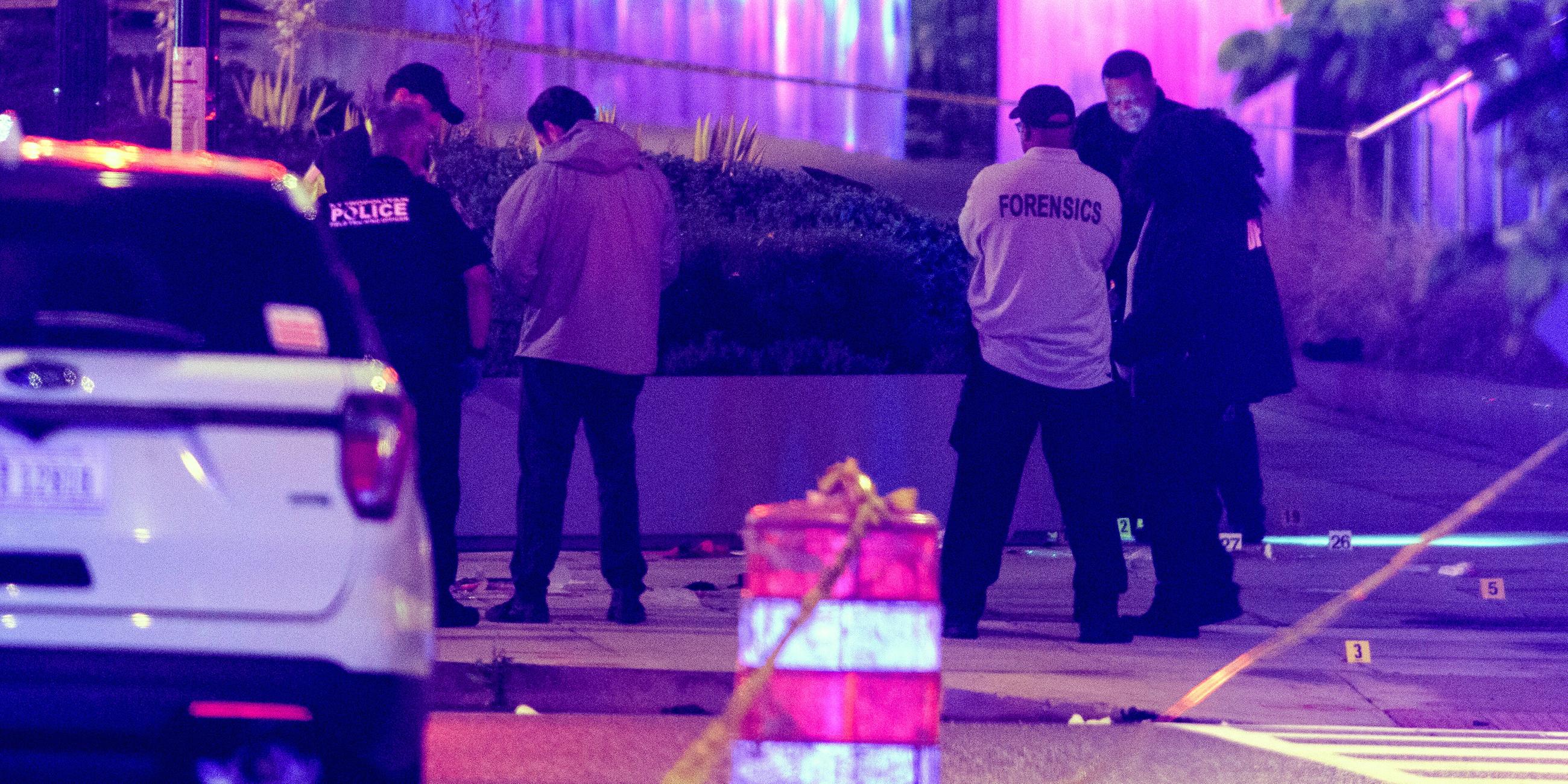 Attentatszenerie nach dem Anschlag, links ein weißes Auto von hinten, aus der Ferne die Ermittler/Polizisten von hinten