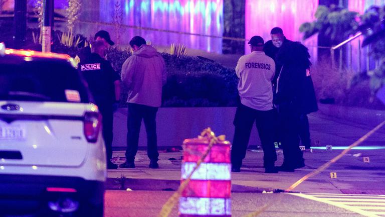 Attentatszenerie nach dem Anschlag, links ein weißes Auto von hinten, aus der Ferne die Ermittler/Polizisten von hinten