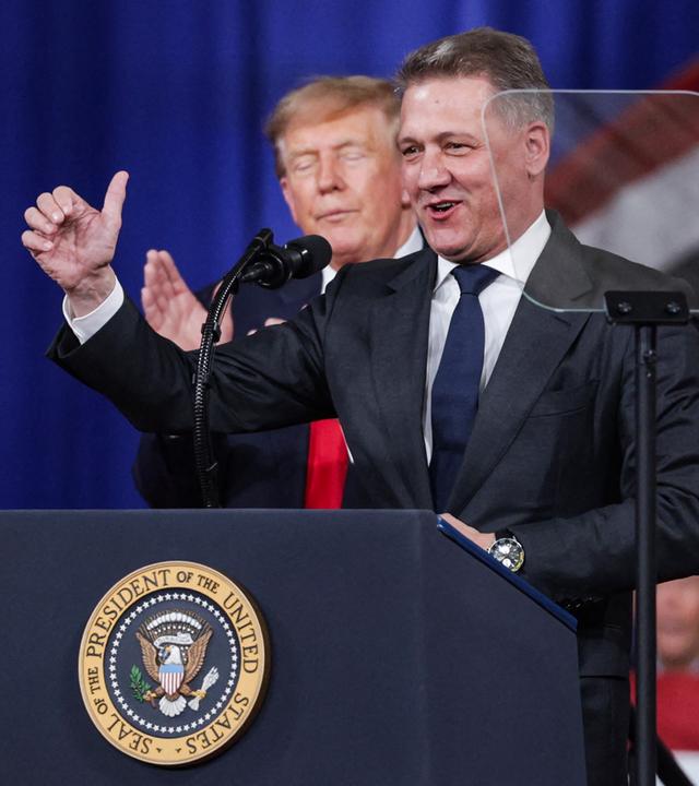 Audi-Chef Döllner steht auf einer Bühne am Rednerpult mit Mikrofon, hinter ihm Donald Trump, der einen Daumen in die Höhe reckt.