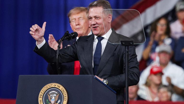 Audi-Chef Döllner steht auf einer Bühne am Rednerpult mit Mikrofon, hinter ihm Donald Trump, der einen Daumen in die Höhe reckt.
