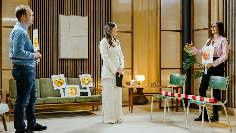 Personalerin Ivana Tadic hält ein Schild mit einem wütenden Smiley hoch. Sie diskutiert mit Dr. Robin Kiera, der ihr gegenüber steht. Psychologin und Host Muriel Böttger steht zwischen beiden und leitet das Gespräch an.