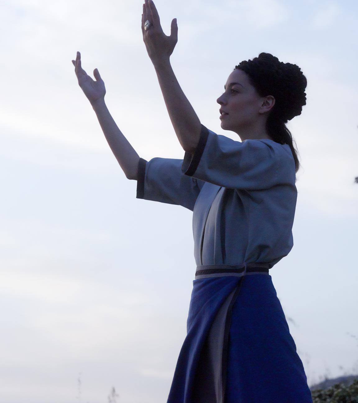 Spielszene: Sijama (Rania Athanasopoulou) steht am Heiligen Berg und spricht ein Gebet. Sie hat ihre Hände gen Himmel erhoben.