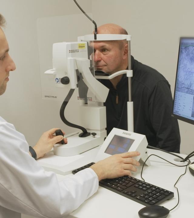 Mann und Arzt sitzen sich gegenüber; Mann schaut mit einem Auge in ein medizinisches Gerät
