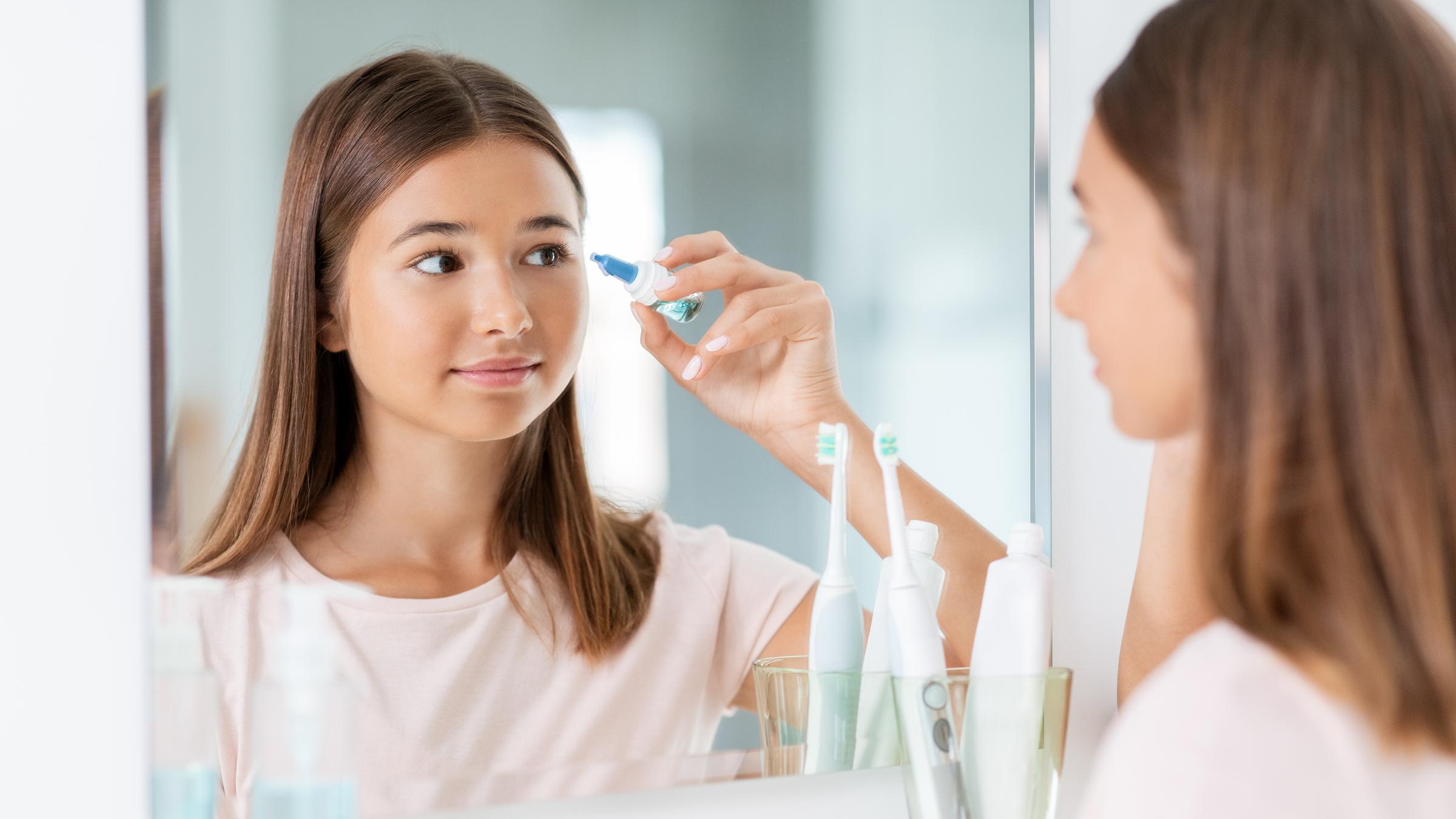 Ein Mädchen steht in einem Badezimmer vor dem Spiegel und verwendet Augentropfen.
