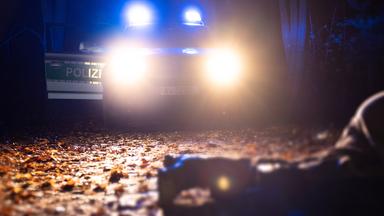 Die Beine eines am Boden liegenden Menschen liegen im Scheinwerferlicht eines Polizeifahrzeugs in der Dunkelheit auf Laub.