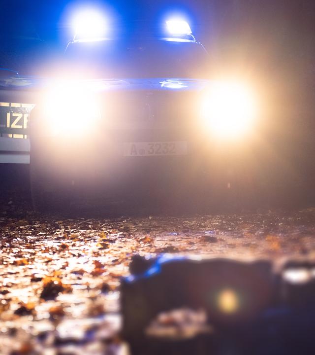 Die Beine eines am Boden liegenden Menschen liegen im Scheinwerferlicht eines Polizeifahrzeugs in der Dunkelheit auf Laub.