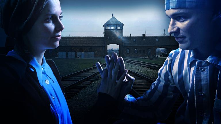 Im Vordergrund ein Liebespaar, das sich gegenüber sitzt und die Hände in Verbundeheit gegeneinander hält. Dahinter die Einfahrt zum KZ Ausschwitz