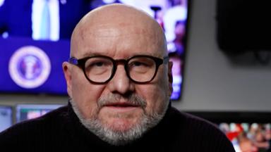 ZDF-Korrespondent Ulf Röller sitzt auf einem Stuhl. Er trägt eine Brille und trägt einen dunklen Pullover. Im Hintergrund leuchtet ein Bildschirm, auf dem verschwommen Donald Trump zu sehen ist.