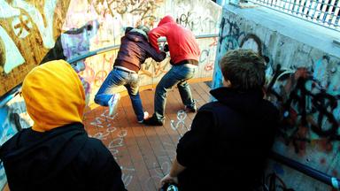Zwei Jungen, einer mit roter und einer mit brauner Jacke, sind in eine Prügelei verwickelt. Beide tragen Kapuzen, ihre Gesichter sind nicht zu sehen. Zwei weitere Jugendliche schauen zu.