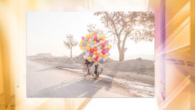 In einer staubigen Landschaft sieht man ein Fahrrad, welches von vielen, bunten Luftballons verdeckt ist.