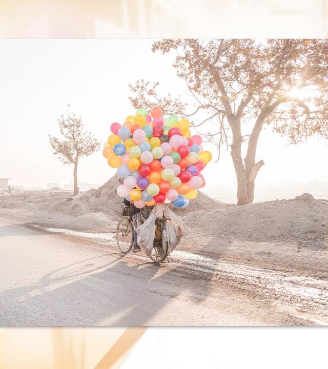 In einer staubigen Landschaft sieht man ein Fahrrad, welches von vielen, bunten Luftballons verdeckt ist.