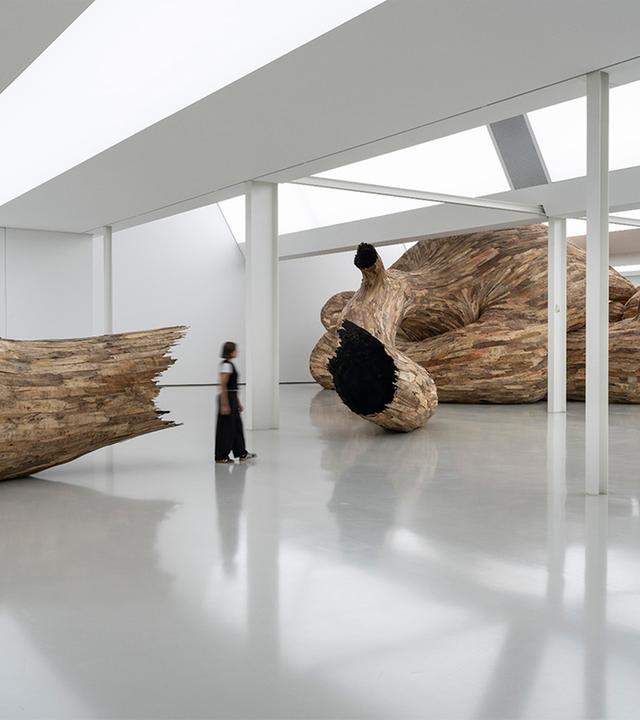 Eine Person steht in einem Ausstellungsraum, in dem die großformatige Installation eines hölzernen Kunstwerkes zu sehen ist.