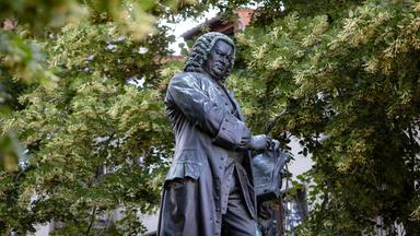 Statue des Komponisten Johann Sebastian Bach - man sieht die Skulptur eines Mannes in historischem Gehrock vor grünen Bäumen vor einem Gebäude.