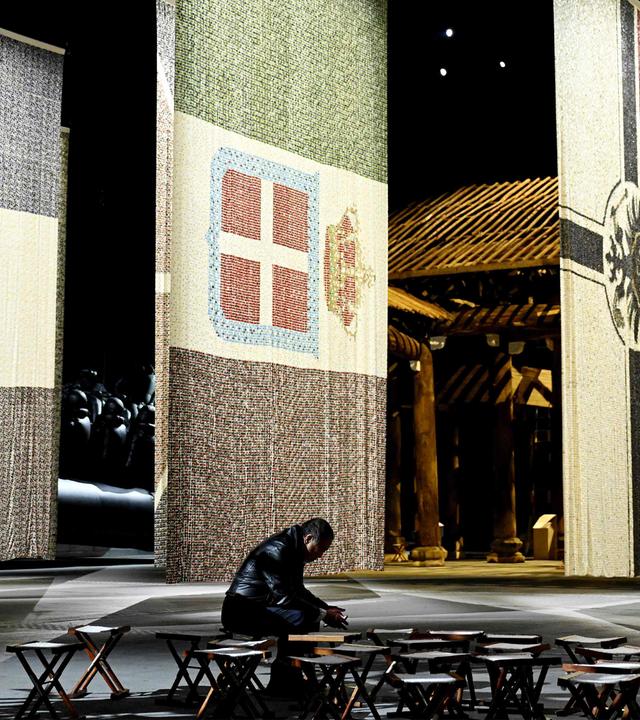 In einem großen dunklen Ausstellungsraum sind riesige Flaggen aus Knöpfen hintereinander ausgestellt. Im Vordergrund ist der Künstler klein und undeutlich zu sehen, wie er auf einem Stuhl in einer Gruppe aus Stühlen sitzt.