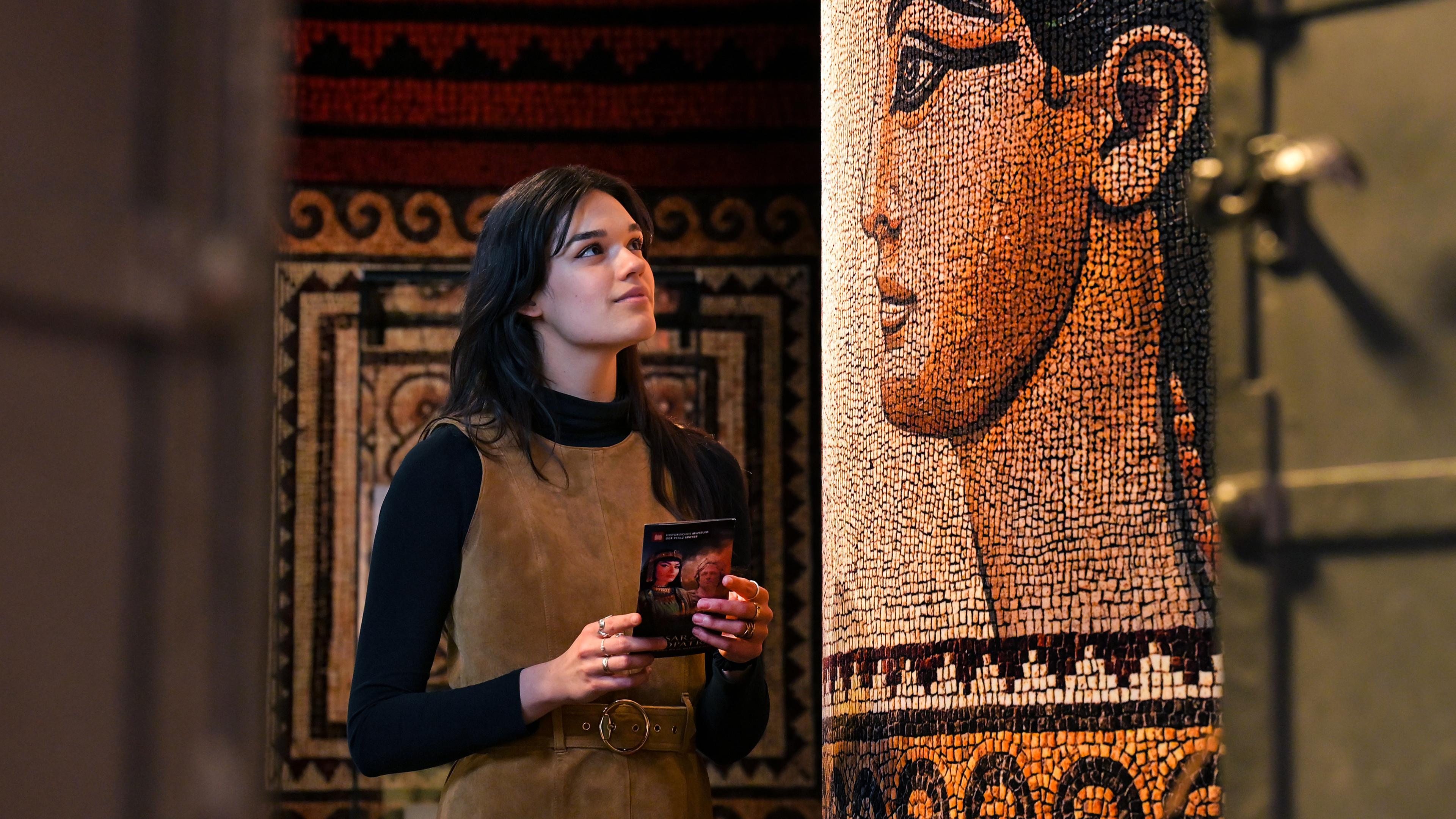 In einem Ausstellungsraum schaut sich eine junge, dunkelhaarige Frau ein Mosaik eines Gesichtes einer Frau aus dem alten Ägypten an.