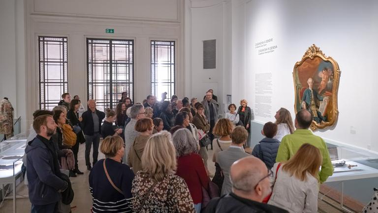 In einem Ausstellungsraum schauen sich viele Personen ein altes Gemälde rechts an der Wand an.