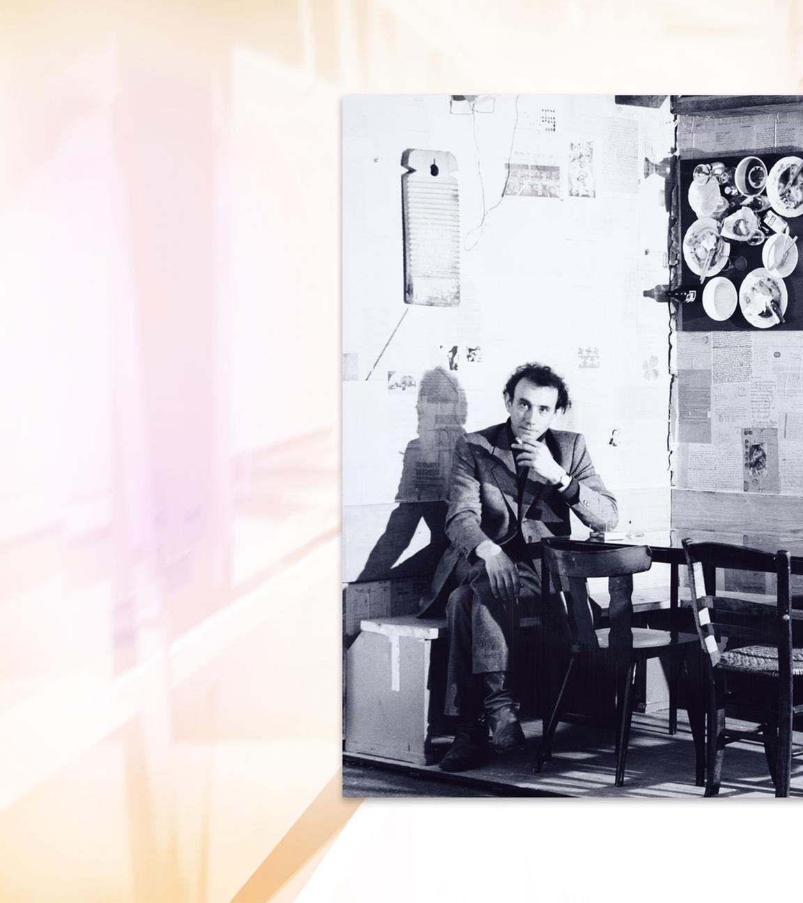 Auf einer Schwarz-Weiß-Fotografie sitzt ein Mann auf einer Bank in einer Ecke. An den Wänden hängt Geschirr.