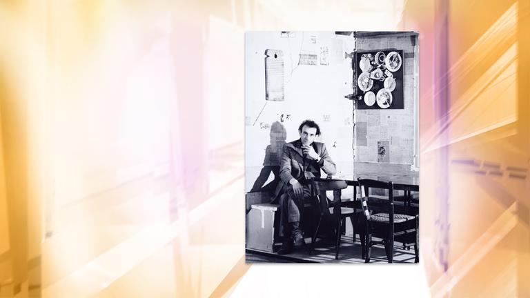 Auf einer Schwarz-Weiß-Fotografie sitzt ein Mann auf einer Bank in einer Ecke. An den Wänden hängt Geschirr.