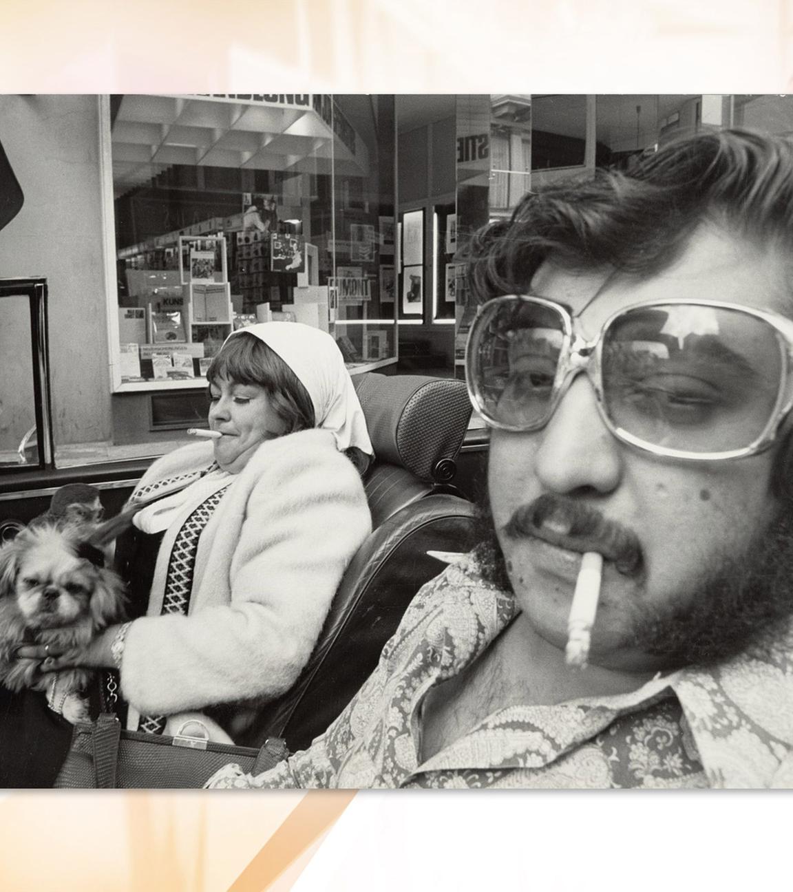 Eine Frau mit Hund auf dem Schoß und ein Mann sitzen rauchend in einem Cabrio. Sie tragen Kleidung der 1970er Jahre.
