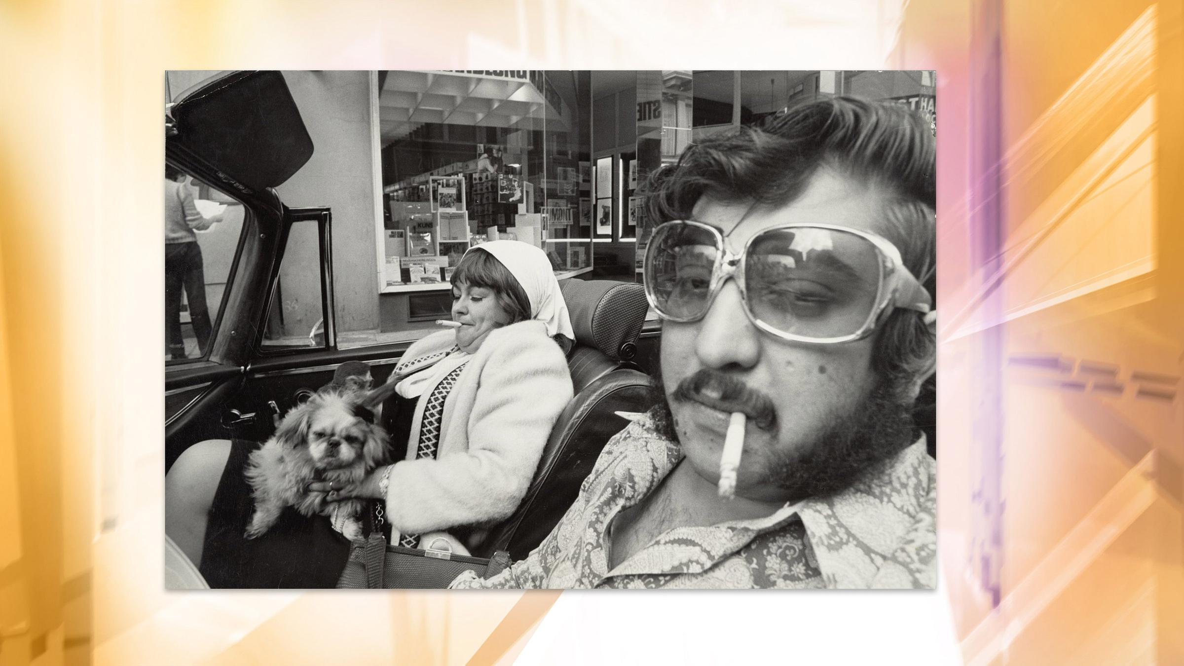 Eine Frau mit Hund auf dem Schoß und ein Mann sitzen rauchend in einem Cabrio. Sie tragen Kleidung der 1970er Jahre.