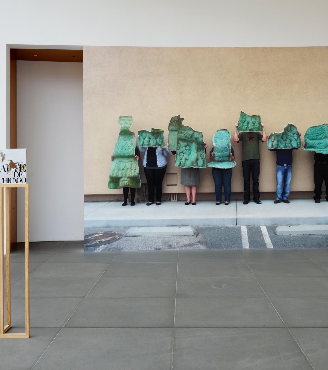 In einem Austellungsraum stehen Exponate und an der Wand befindet sich eine große Fotografe, die Menschen zeigt, die grüne Gegenstände vor sich halten.