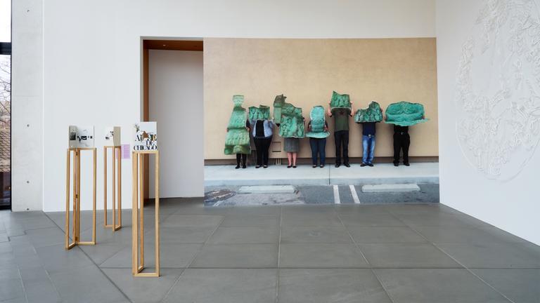 In einem Austellungsraum stehen Exponate und an der Wand befindet sich eine große Fotografe, die Menschen zeigt, die grüne Gegenstände vor sich halten.