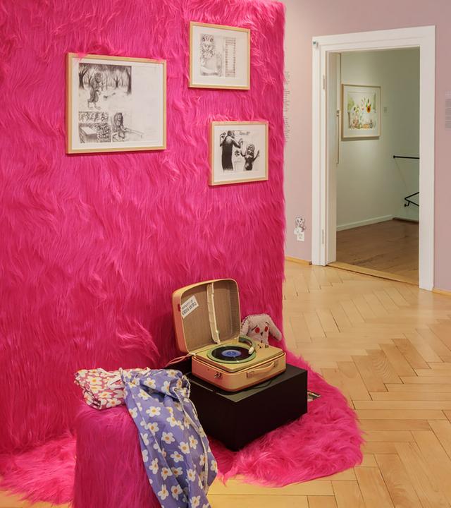 In einem Ausstellungsraum sieht man Illustrationen an einer mit pinkfarbenem Zottelstoff verkleideten Wand hängen. Im Vordergrund steht ein altmodischer Plattenspieler.