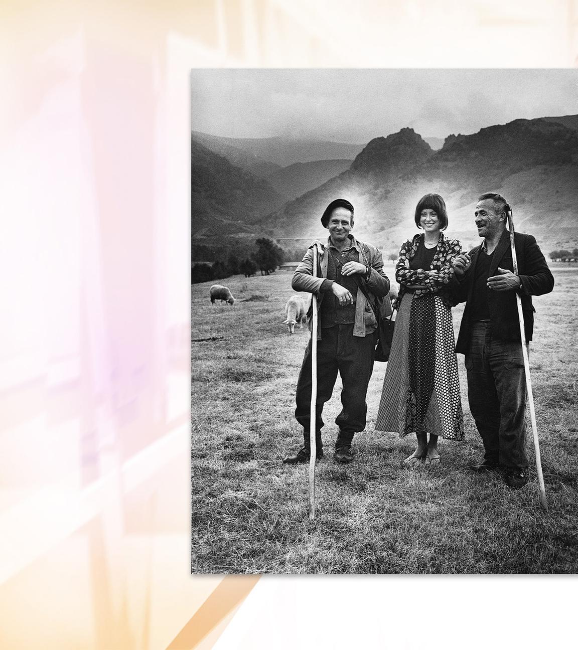 Auf einer Schwarz-Weiß-Fotografie sieht man eine Frau, die zwischen zwei Schäfern mit langen Stöcken auf einer Wiese vor hügeliger Landschaft steht.