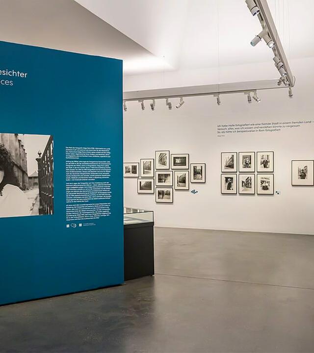 In einem Ausstellungsraum sieht man gerahmte Schwarz-Weiß-Fotografien an den Wänden hängen. Im Raum steht eine groß Stellwand mit einem vergrößerten Foto und Infos dazu.