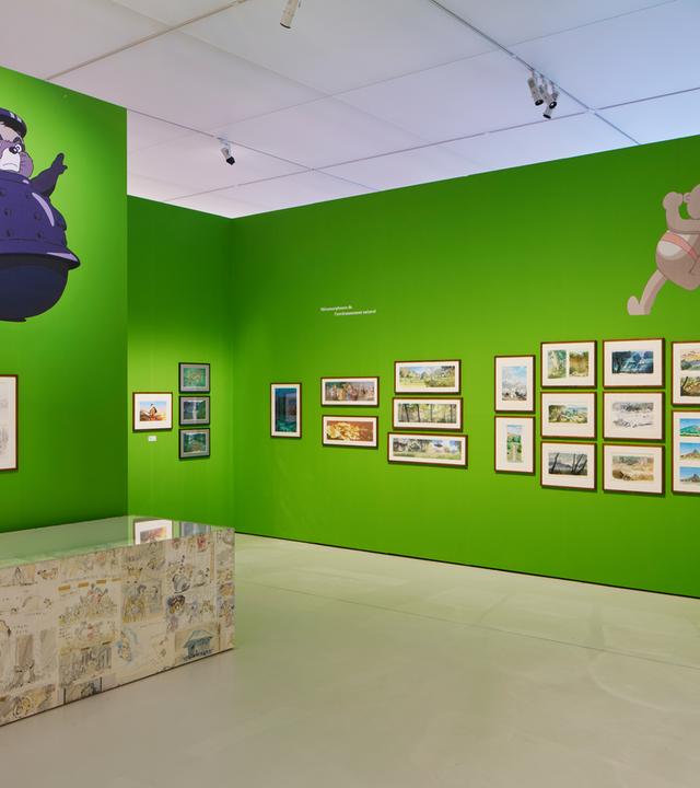 Ausstellungsansicht: Man sieht Bilder an einer grünen Wand hängen und Comic-Zeichnungen von Tieren auf den Wänden.